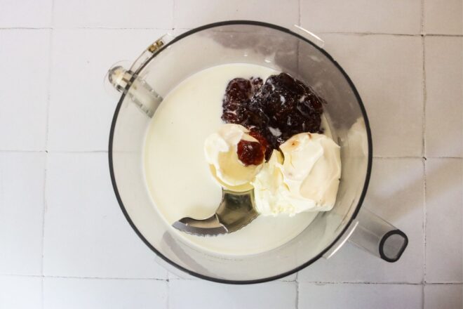 This is an overhead horizontal image of a food processor with cream cheese, milk and strawberry jam in it. The food processor sits on a white square tile surface.