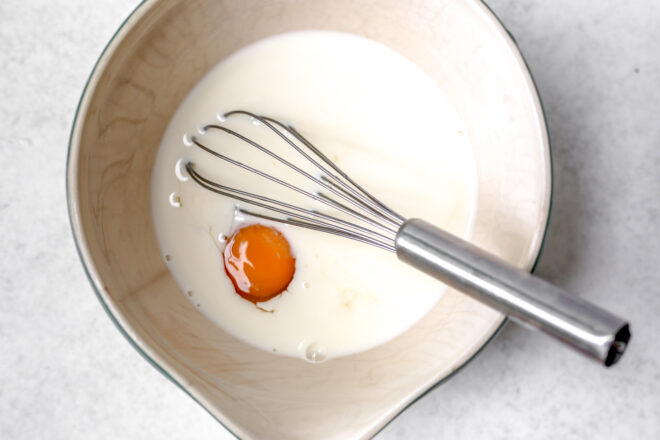 This is an overhead horizontal image of a large beige bowl with a spout. I the bowl is milk and an egg. A silver whisk is in the bowl with the handle leaning against the rim of the bowl pointing to the bottom right corner of the image. 