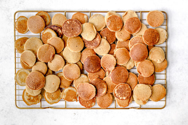 This is an overhead horizontal image of a gold cooling rack with a lot of mini pancaked piled on top of each other. The cooling rack sits on a white surface. 