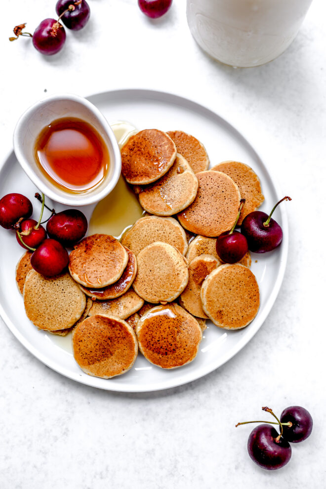 This is an overhead vertical image of a large white plate with a lot of mini pancakes on it. A small white bowl with syrup is also on the plate and has been drizzled onto the pancakes. Cherries are on the plate with the pancakes and some are on the white surface above and below the plate. A glass jug with milk is in the top right corner of the image. 