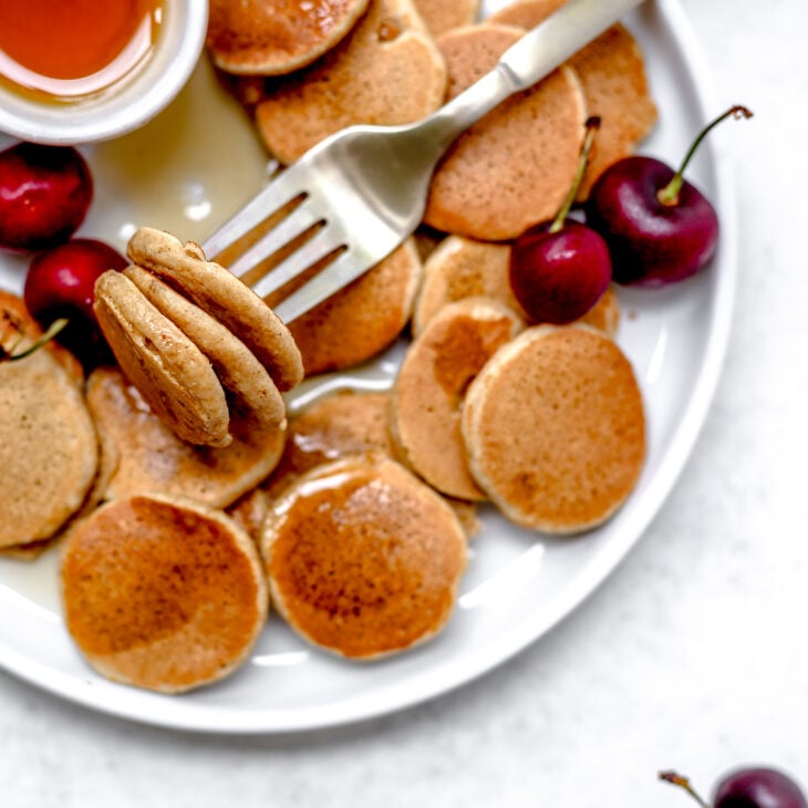 This is an overhead horizontal image of a white plate with mini pancakes on it. A fork is piercing three pancakes and leaning on the plate on top of the rest of the pancakes. A small white bowl with syrup is on the plate and has been drizzled on top of the pancakes. Cherries are on the plate and one cherry is off the plate in the bottom right corner on the white surface.