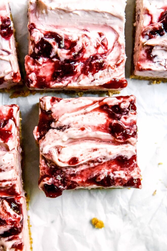 This is an overhead vertical image of a light pink cheesecake square with swirls of cherry jam. The square is in the center of the image with more squares on the white surface above and to the left of the image.