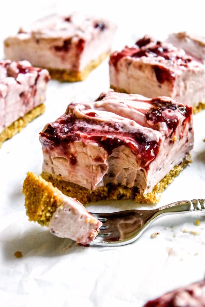 This is a vertical image looking at a cherry cheesecake square from the side. The cheesecake is light pink, has a graham cracker crust, and swirls of cherry jam on top. A silver fork has a bite of cheesecake on it and lies on the white surface in front of the cheesecake square. More cherry cheesecake squares are blurred behind and to the side of the slice in focus.