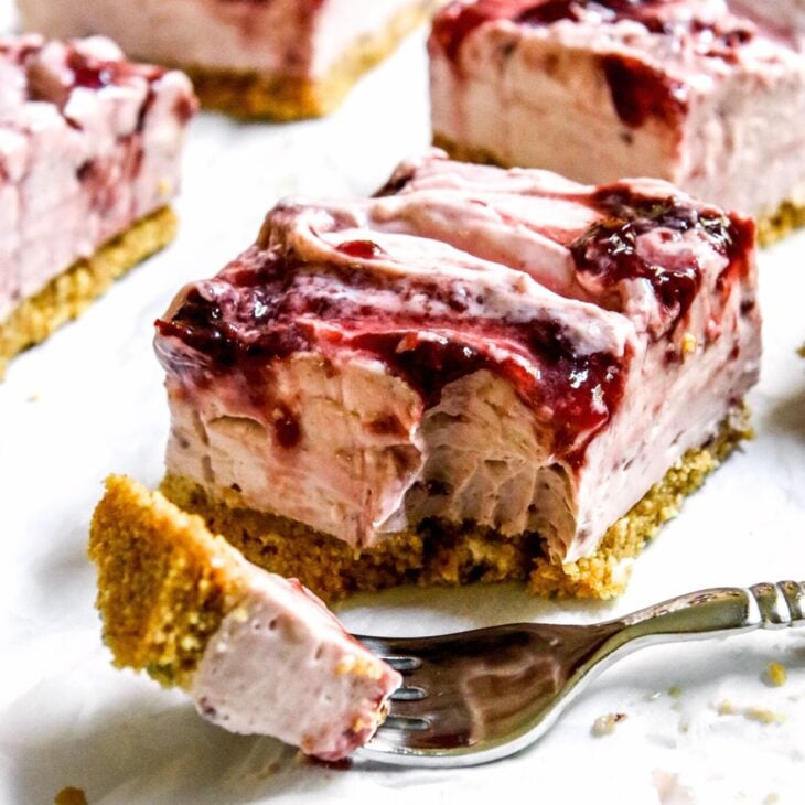 This is a vertical image looking at a cherry cheesecake square from the side. The cheesecake is light pink, has a graham cracker crust, and swirls of cherry jam on top. A silver fork has a bite of cheesecake on it and lies on the white surface in front of the cheesecake square. More cherry cheesecake squares are blurred behind and to the side of the slice in focus.