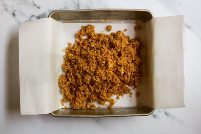 This is an overhead horizontal image looking down into a square metal pan on a white marble surface. The pan is lined with parchment paper and a graham cracker crust is in a pile on top.