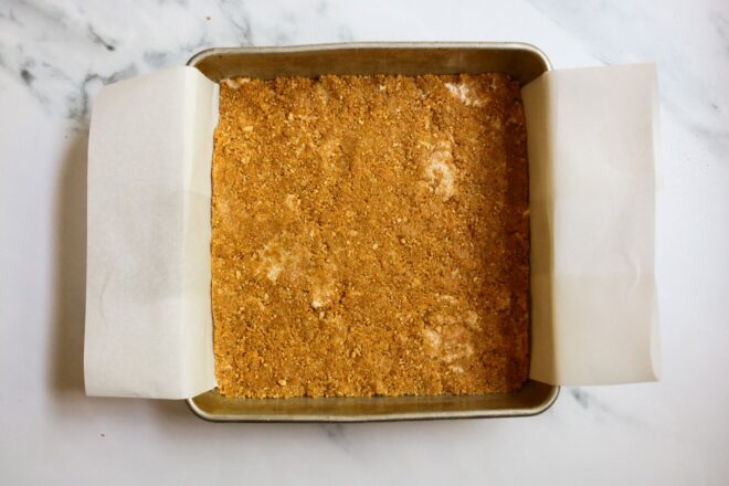 This is an overhead horizontal image looking down into a square metal pan on a white marble surface. The pan is lined with parchment paper and a graham cracker crust is pressed into the bottom.