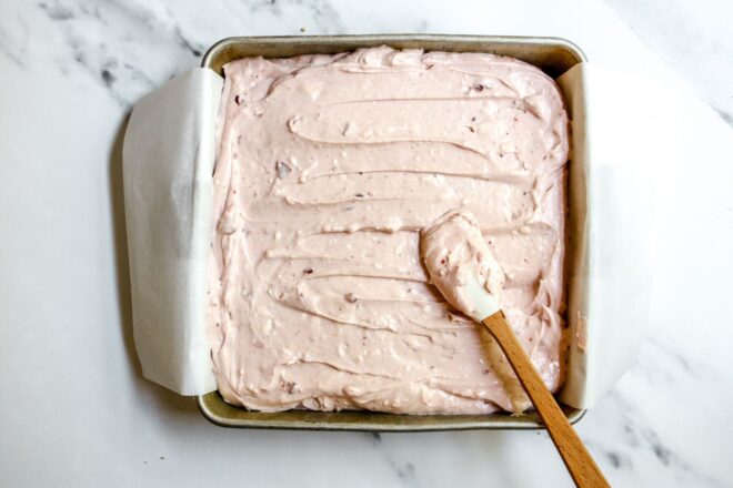 This is an overhead horizontal image looking down into a square metal pan on a white marble surface. In the pan is a light pink cream cheese mixture spread out with a small white and wood spatula leaning against the bottom right corner of the pan.