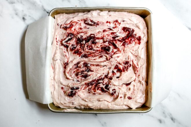 This is an overhead horizontal image looking down into a square metal pan on a white marble surface. In the pan is a ligh pink cream cheese mixture spread out with swirls of cherry jam on top.