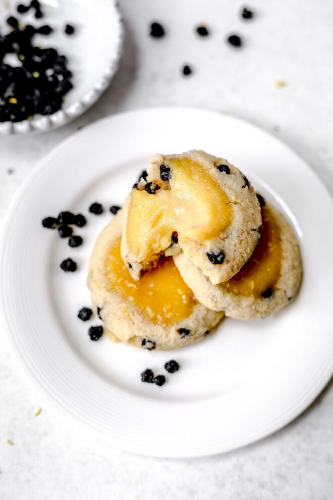This is vertical image looking a small white plate with three blueberry cookies with lemon curd in the middle. The plate sits on a light grey surface with a small bowl of dried blueberries in the back left corner of the image. More dried blueberries are scattered around the plate and surface.