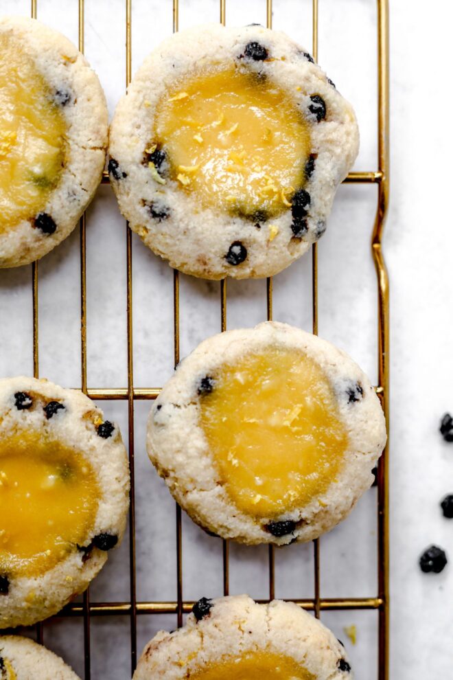 This is an overhead vertical image of a gold cooling rack with cookies on it. The cookies are studded with blueberries and have a lemon curd pool in the middle. The cooling rack is in a light grey surface.