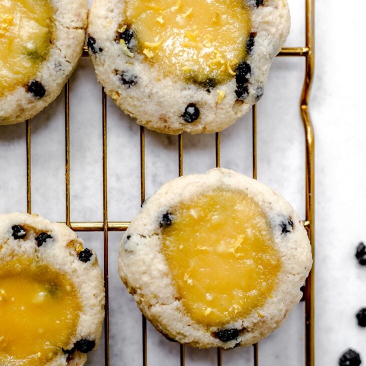 This is an overhead vertical image of a gold cooling rack with cookies on it. The cookies are studded with blueberries and have a lemon curd pool in the middle. The cooling rack is in a light grey surface.