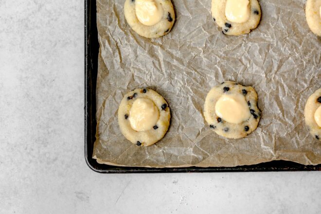 This is and overhead horizontal image of a rimmed baking sheet lined with a crumpled brown piece of parchment paper. On the parchment paper are six flattened blueberry cookies with a lemon curd puddle in the center. The baking sheet sits on a light grey surface.