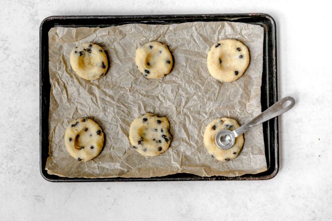 This is and overhead horizontal image of a rimmed baking sheet lined with a crumpled brown piece of parchment paper. On the parchment paper are six flattened blueberry cookies with dimples in the middle of the cookie. A small measuring spoon is in the bottom right corner cookie pressing the back into the top. The baking sheet sits on a light grey surface.