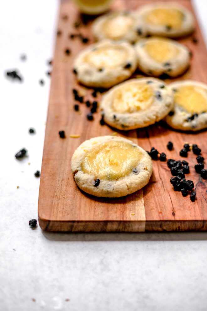 This is a vertical image looking at a wooden serving platter on a light grey surface. On the rectangular platter are lemon blueberry cookies with a lemon curd in the center. Dried blueberries are scattered on the platter and on the grey surface next to the board.