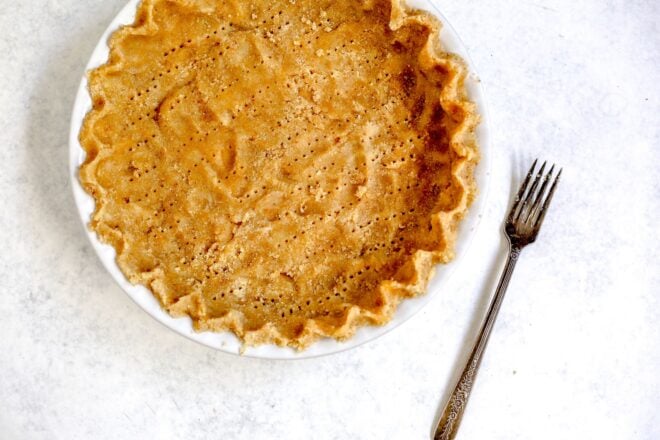 This is a horizontal overhead image looking at a white pie dish. In the pie dish is a light beige colored pie crust with fork holes poked throughout it. The pie crust is crimped and pinched around the edges to create a scalloped edge. The pie dish sits on a light grey and white surface with a fork on the surface to the right side of the pie dish.