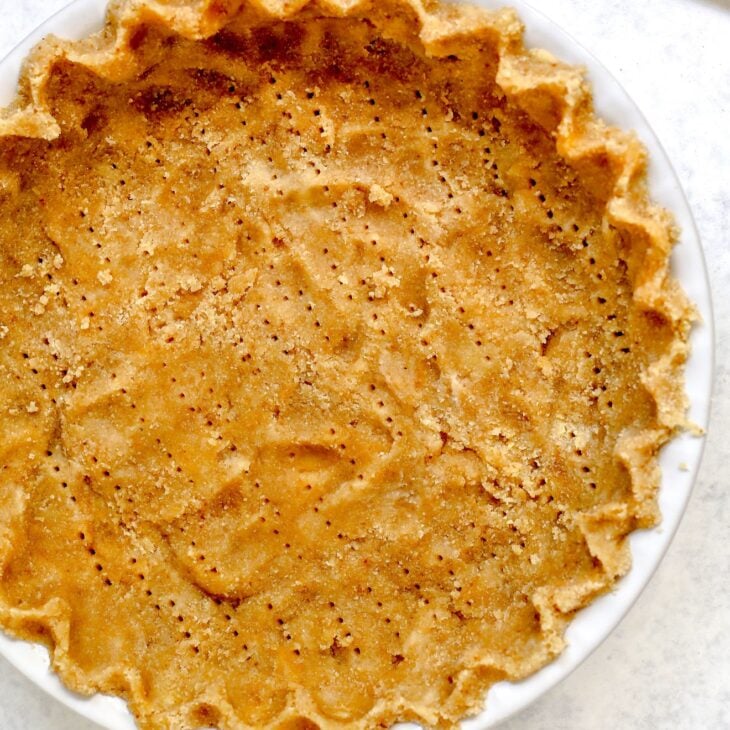 This is an overhead vertical image looking at a white pie dish. In the pie dish is a light beige colored pie crust with fork holes poked throughout it. The pie crust is crimped and pinched around the edges to create a scalloped edge. The pie dish sits on a light grey and white surface.