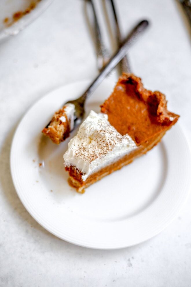 This is an overhead vertical image of a small white plate with a slice of pumpkin pie on it. The pie has a dollop of whipped cream and a sprinkle of ground cinnamon. The plate sits on a light grey, almost white surface and a fork, with a bite of pumpkin pie on it, is on the plate with the handle leaning against the edge of the plate pointing to the top right corner of the image.