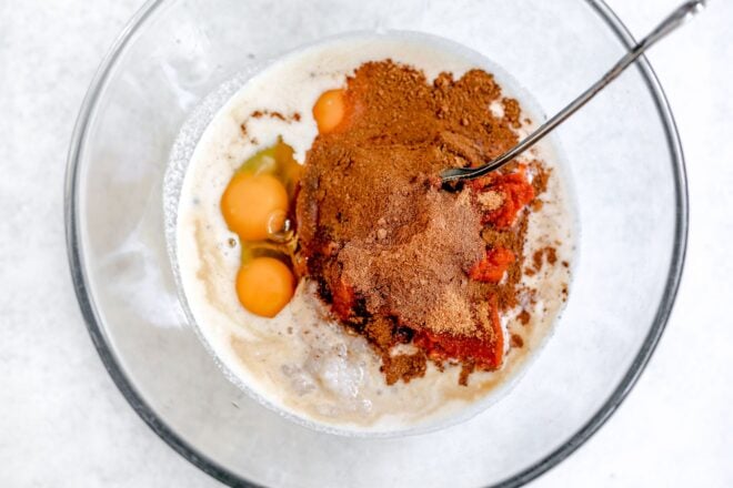 This is an overhead horizontal image of a glass bowl with eggs, milk, coconut sugar, and pumpkin puree in it. A silver utensil is dipping into the bowl with the handle leaning against the side of the bowl, pointing to the top right corner of the image. The bowl sits on a light grey surface.