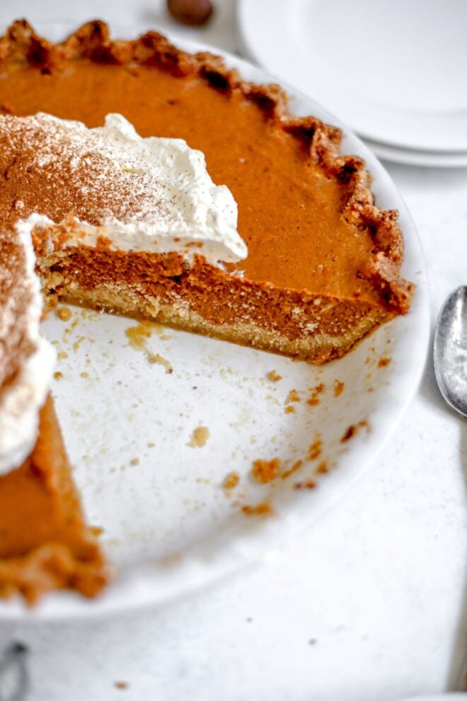 This is a vertical image looking at a pumpkin pie from an overhead angle. The pumpkin pie is cut into with slices taken out and the image focuses on the layers of crust and pumpkin. The pie has a large dollop of whipped cream and a sprinkle of ground cinnamon. The pie dish sits on a white surface with a small stack of white plates to the top right corner and a spoon peeking in from the right side of the image.