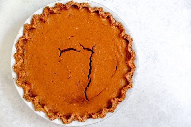 This is an overhead horizontal image of a white pie dish with a light brown pinched pie crust with baked pumpkin pie filling in it. The surface of the pumpkin pie has cracks ion it. The pie dish sits on a light grey, almost white surface.