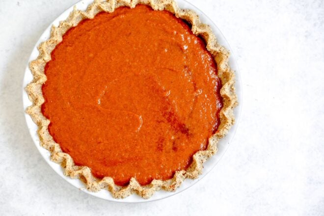 This is an overhead horizontal image of a white pie dish with a light brown pinched pie crust with a raw pumpkin pie filling in it. The pie dish sits on a light grey, almost white surface.