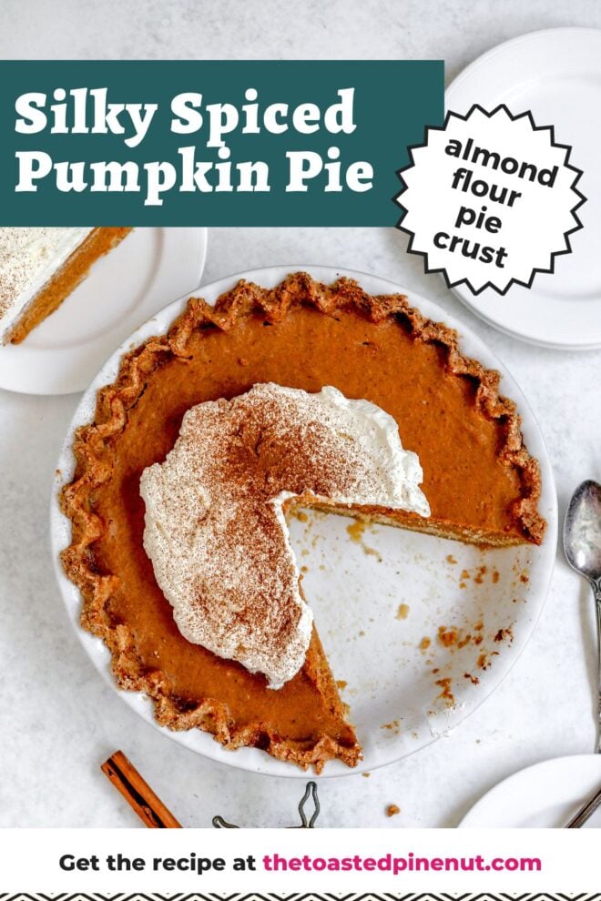 This is an overhead vertical image of a white pie dish with a light brown pinched pie crust with baked pumpkin pie filling in it. On top of the pumpkin pie is large dollop of whipped cream and a sprinkle of ground cinnamon. The pie is sliced into with a large wedge of slices taken out of it. The pie dish sits on a light grey, almost white surface. Antique silverware is to the right side of the pie dish. To the top right corner of the image is a couple small white plates stacked on top of each other. To the top left is a plate with a slice of pumpkin pie and a stick of cinnamon is to the bottom of the image. Text overlay reads "silky spiced pumpkin pie almond flour pie crust" at the top and "get the recipe at thetoastedpinenut.com" at the bottom of the image.