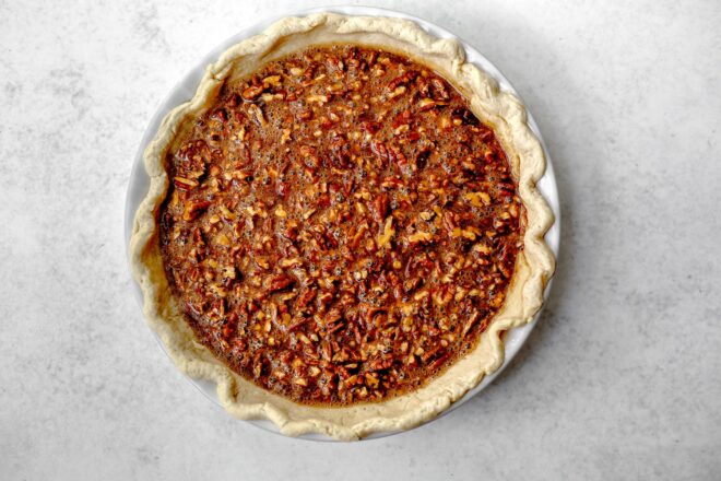 This is an overhead horizontal image of a white pie dish with a pecan pie in it before it's been baked. The pie dish sits on a light grey surface.