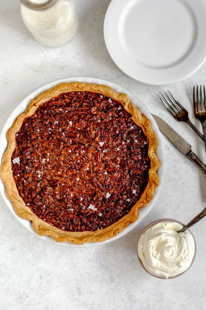 This is an overhead vertical image of a white pie dish with a pecan pie in it. The pie dish is on a light grey surface with a small bowl of whipped cream with a spoon in it. To the right of the pie dish is two forks and a knife. To the top of the image is a small white plate and a glass jar of milk is in the top left corner of it.