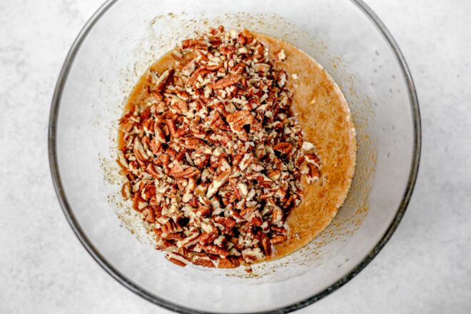 This is an overhead horizontal image of a large glass bowl with a light brown mixture with chopped pecans on top. The bowl sits on a light grey surface.