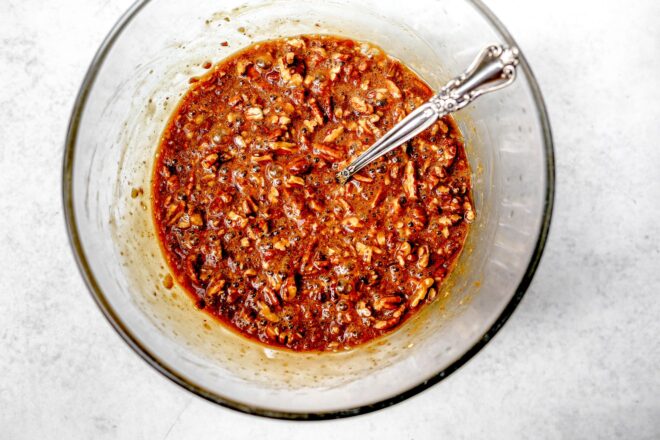 This is an overhead horizontal image of a large glass bowl on a light grey surface. In the bowl is a brown liquid base with chopped pecans in it. A silver utensil is in the mixture with the handle pointing to the top right corner of the image.