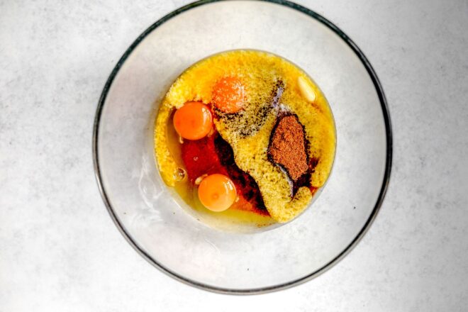 This is an overhead horizontal image of a glass bowl on a light grey surface. In the bowl is eggs, brown sugar, eggs, and vanilla.