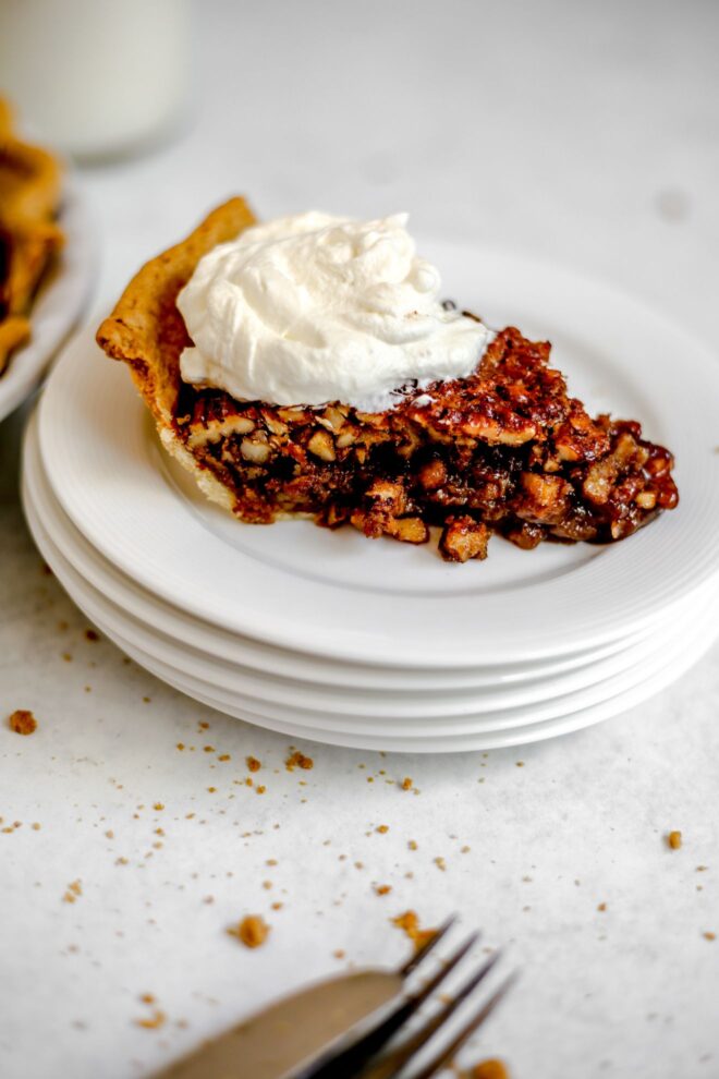 This is a vertical image looking at stack of small white plates with a slice of pecan pie on it. The slice of pie is topped with a dollop of whipped cream. The plates sit on a light grey surface with some crumbs scattered around it. Blurred in the front, bottom of the image are a fork and knife. To the top left corner is a pie and glass of milk peeking in from the side.