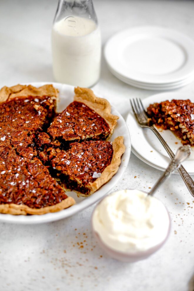 This is a vertical image looking at a white pie dish with sliced pecan pie in it from the left side. The pie dish is to the left side of the image on a light grey surface with a small bowl of whipped cream with a spoon in it is in the bottom right corner of the image. To the right of the pie dish are a stack of two plates with a slice of pecan pie on it. To the top of the image are small white plates and a glass jar of milk is in the top left corner of it.