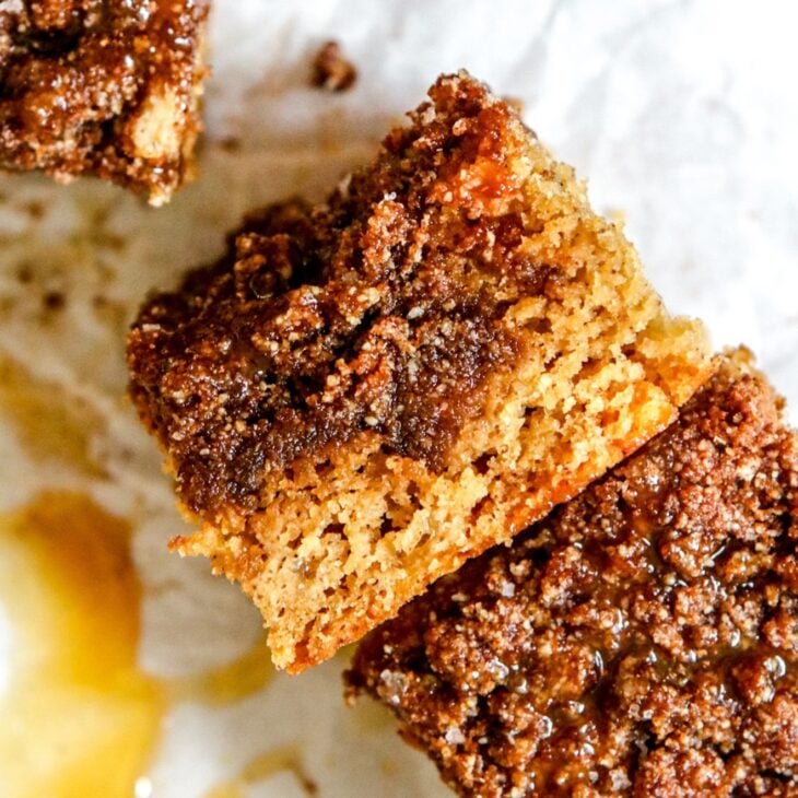 This is a vertical image looking down onto a white piece of parchment paper with a piece of coffee cake leaning on its side so you can see the thick layer of crumble topping and fluffy tender cake. The cake leans against another piece of coffee cake to the bottom right corner of the image. Another piece of coffee cake is in the top left corner of the image. Some caramel and crumbs are on the parchment paper to the bottom left corner of the image.