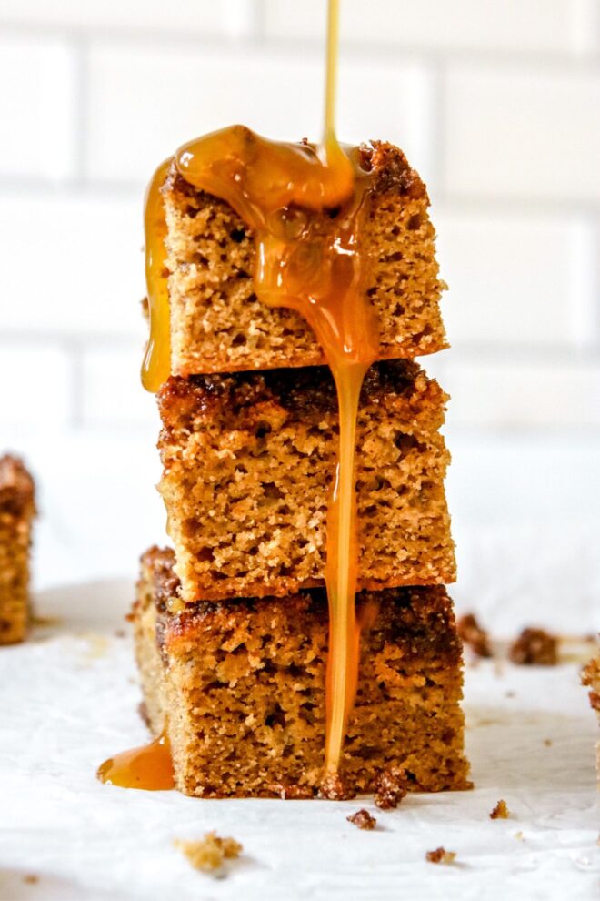 This is a vertical image looking at a stack of three pieces of coffee cake from the side. The stack sits on a white piece of parchment paper with white tile in the background. Caramel is coming in from the top of the image, drizzling on the top of the stack and dripping down the front and sleft side of the stack. Another slice of coffee cake and crumbs are around the stack of coffee cake.