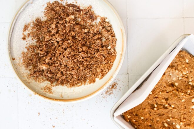 This is an overhead horizontal image of a white shallow bowl with a crumble in it. To the bottom right corner of the image is a square pan lined with parchment paper and a brown batter with white curds throughout it. The bowl and pan sit on a white square tile surface.